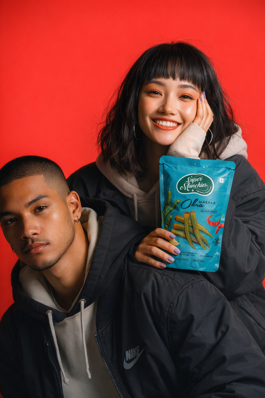 Two people posing with a Super Munchies Masala Okra chips pack against a bold red background, showing a savory snack moment