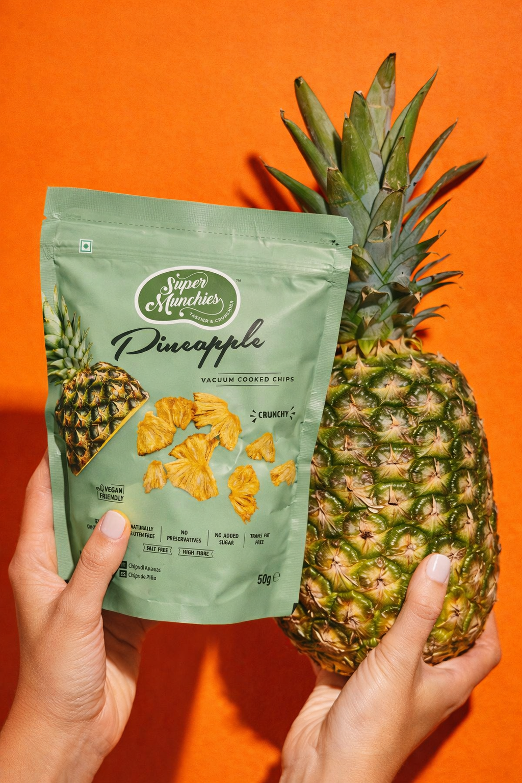 Hands holding a Super Munchies Pineapple chips pack and a whole pineapple, showing a naturally sweet fruit snack on an orange background